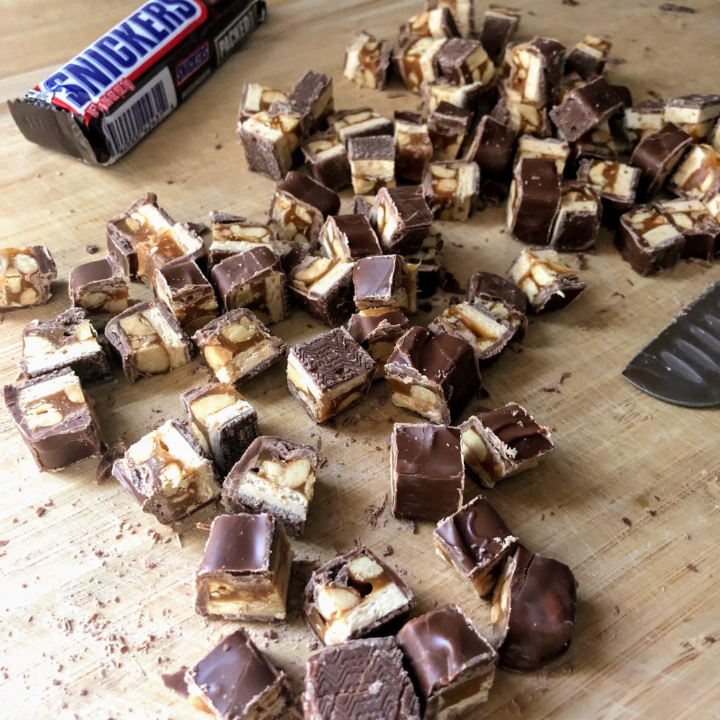 A wooden cutting board has a large knife sitting on it with many chopped pieces of Snickers bars laying on top.