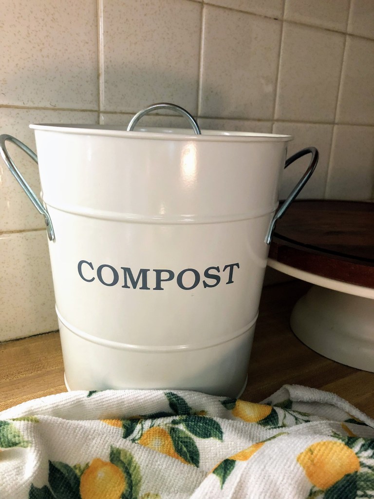 A small compost pail sits on a wood kitchen counter next to a lemon printed kitchen towel.