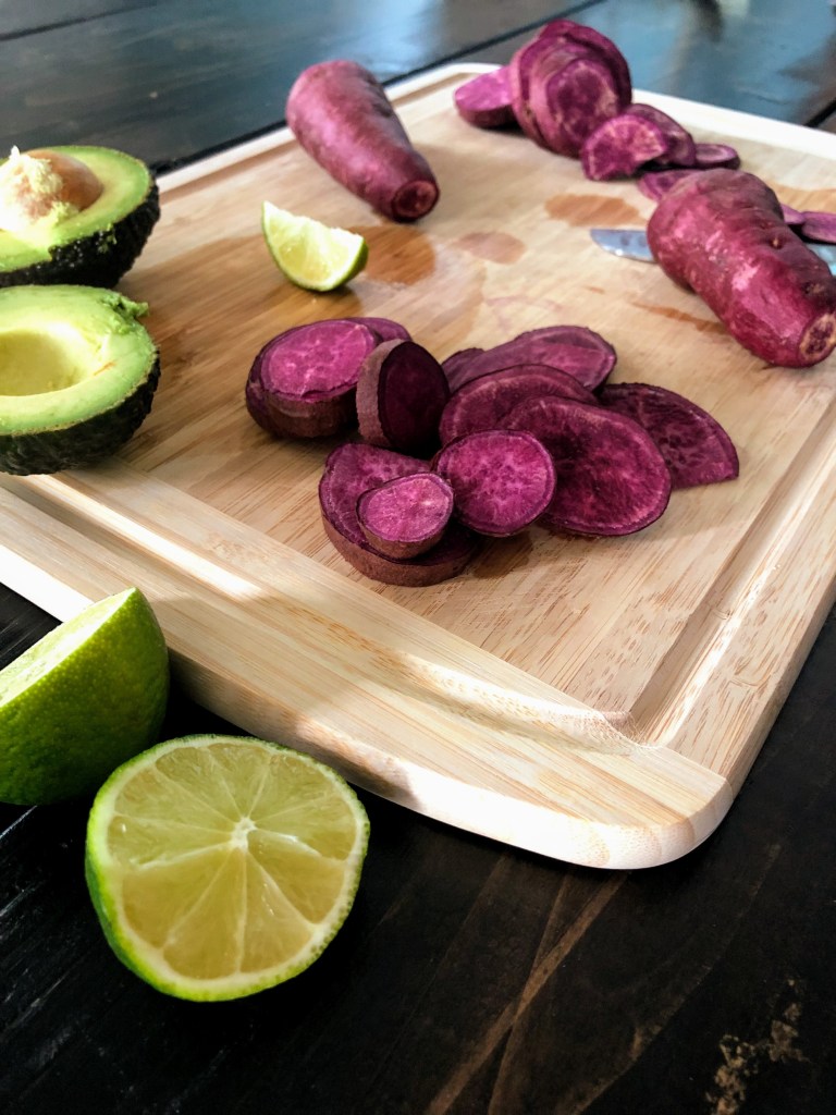Sliced purple sweet potatoes sitting next to avocados on a cutting board.