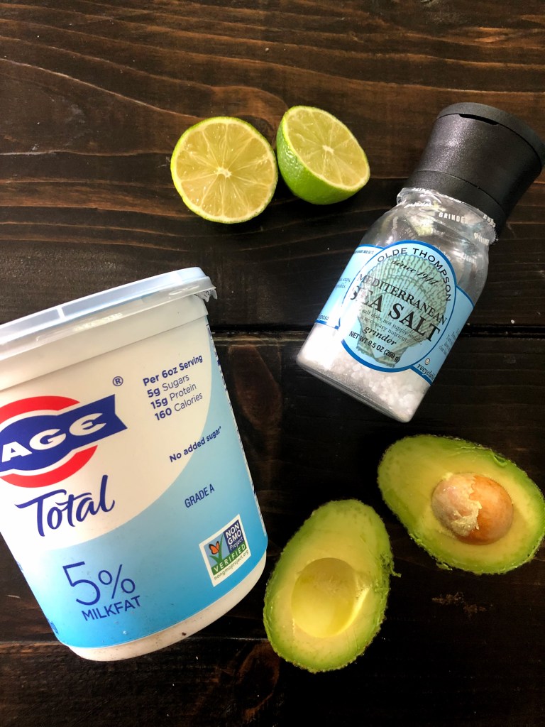 Ingredients consisting of limes, green yogurt, sea salt, and avocados spread across a walnut-stained table.