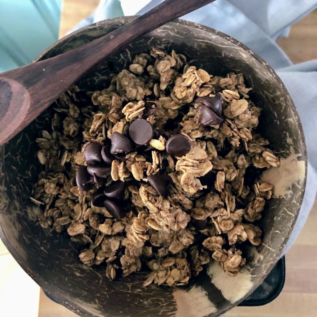 A small wooden bowl of heart healthy, whole grain granola sits atop a linen with a wooden spoon carefully place on the edge of the bowl.