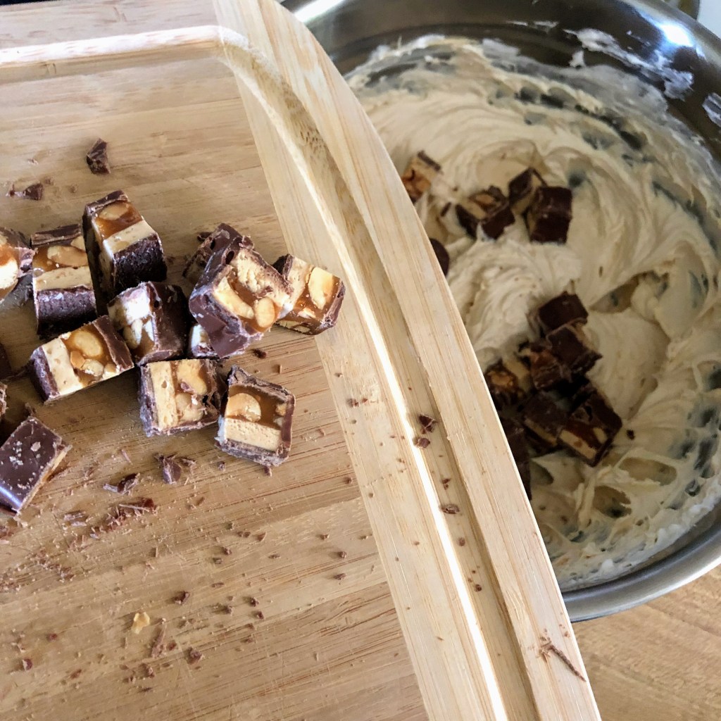 A cutting board with chopped Snickers bars is about to be thrown into a large metal mixing bowl of cream cheese, whipped topping, powdered sugar, vanilla, and peanut butter.