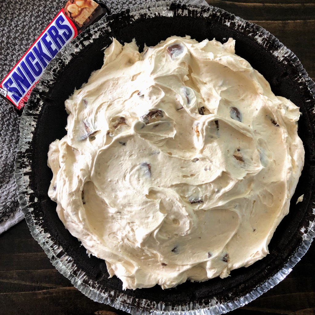 a Snickers pie sits on a grey dish towel with a Snickers bar laying next to it.