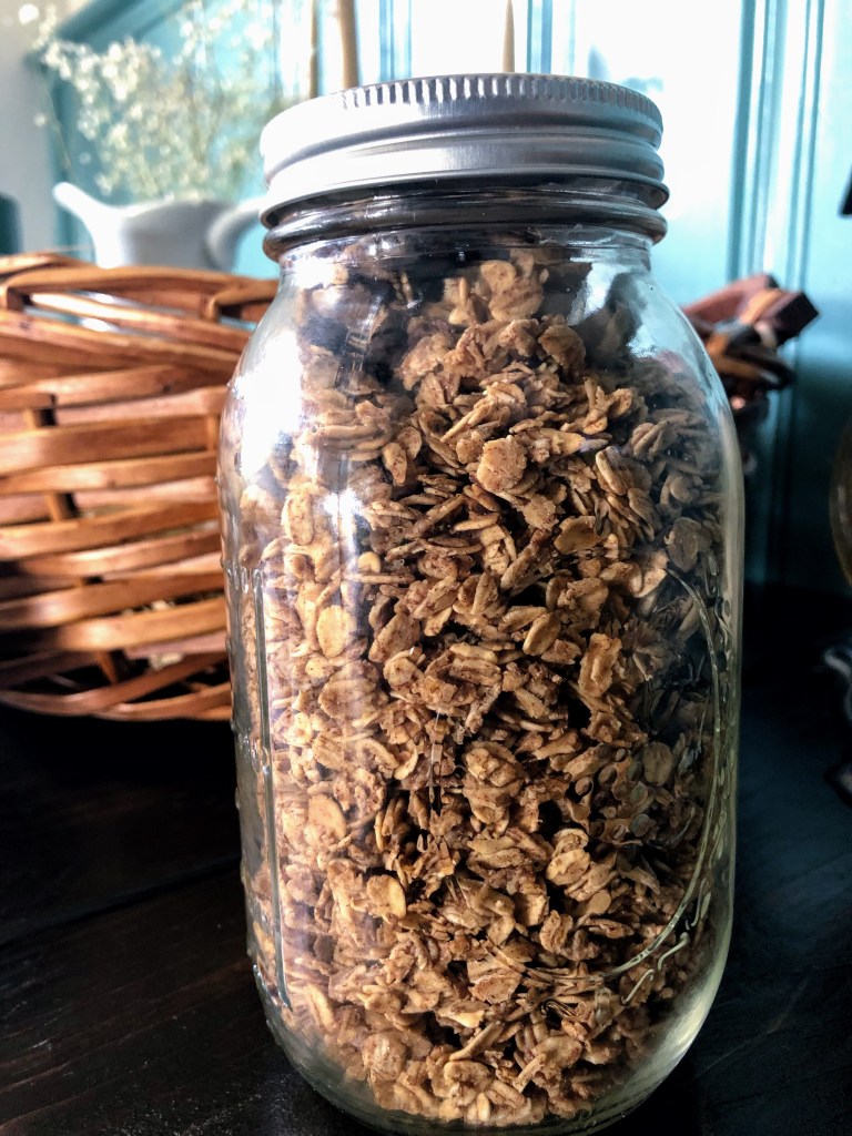 A large glass jar holds homemade heart healthy, whole grain granola in it on a wood dining table with a pitcher of flowers behind it.