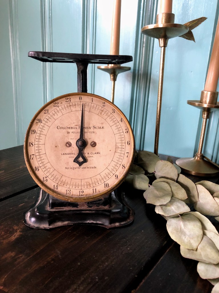 Decorate with antiques with this vintage kitchen scale. It lays next to eucalyptus leaves and  candlestick holders on a dining table.