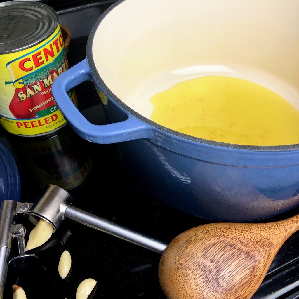 A blue dutch oven is coated in olive oil and is ready for onions and garlic to be thrown in on the stovetop.