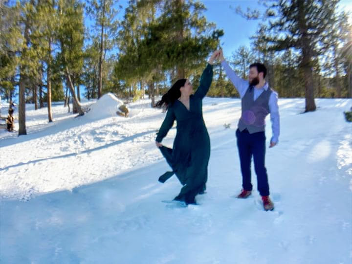 A newly married couple dances in the snow in front of pine trees in a forest.