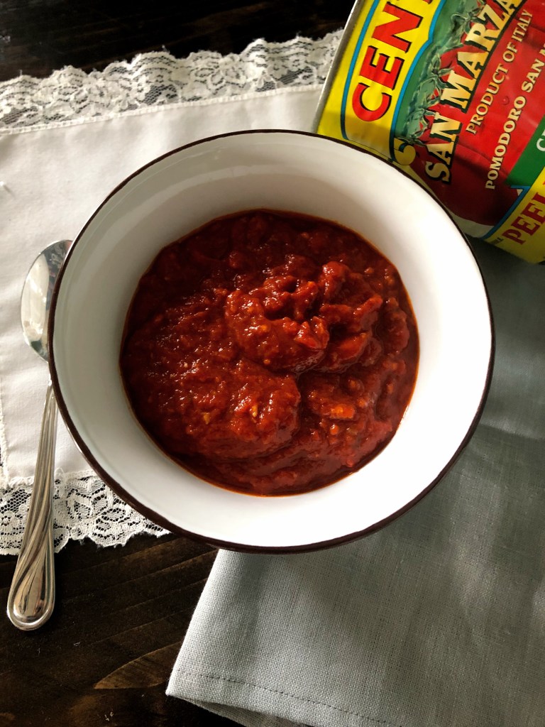 Antique linens sit atop a kitchen table with a can of tomatoes and a bowl of quick and easy marinara.