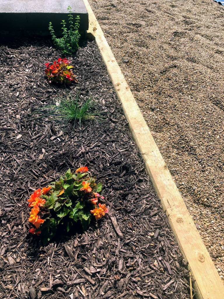 A pea gravel patio is surrounded by timber edging with mulch and plants as landscaping close to a jet black concrete stoop.