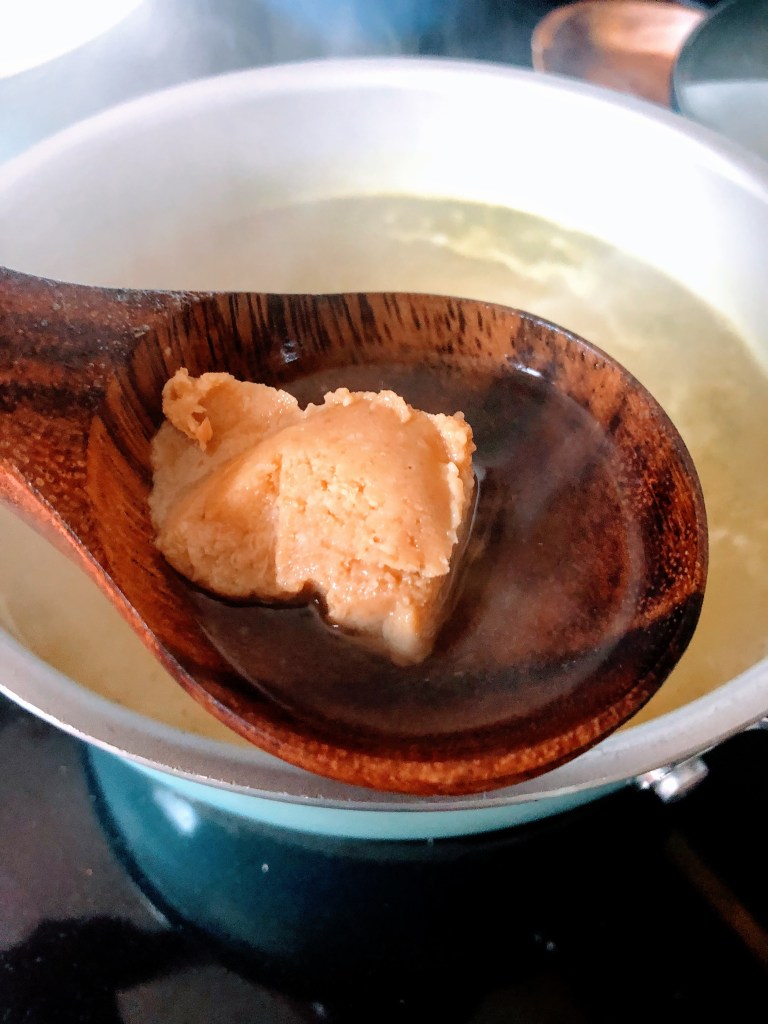 A scoop of white miso is added to a ladle full of boiling water.
