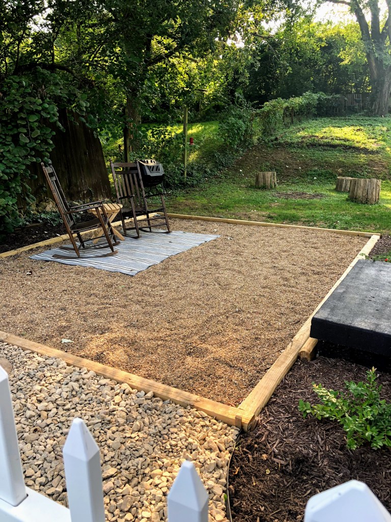 A finished pea gravel patio is lined with unstained timber and has two rocking chairs, a rug, and a small table donning it.