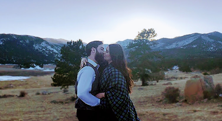 A man and woman kiss in front of the Rocky Mountains.