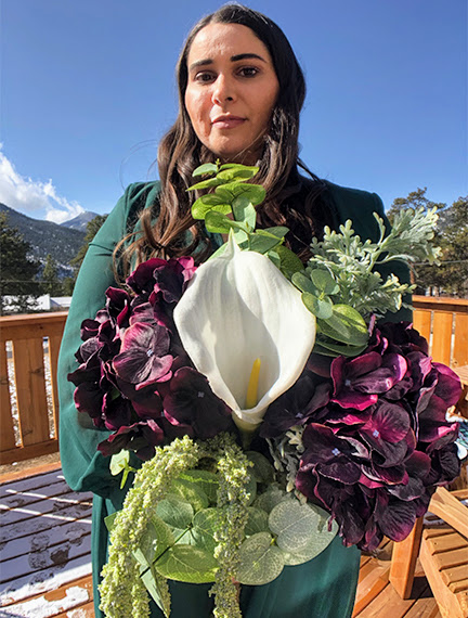 A brunette woman stands in her wedding gown, outside on a cabin deck, with mountains in the background and a bouquet of flowers.