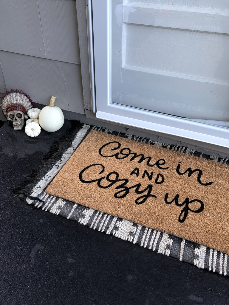 A fall rug that reads “come in and cozy up” sits on a black stoop outside a Midwestern cottage.