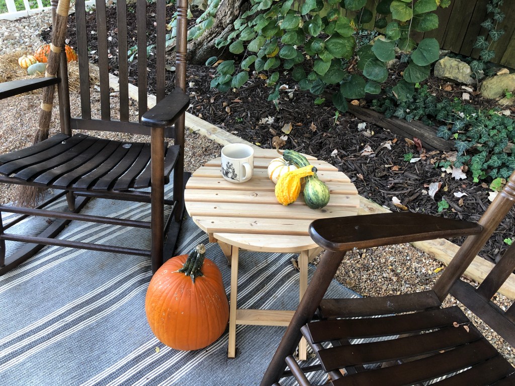Pumpkins and gourds sit on a table next to rocking chairs on a patio.