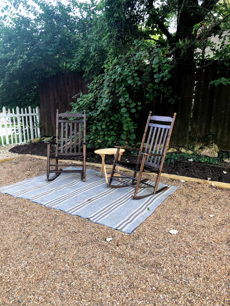 A set of walnut-stained chairs sit above a striped rug on a pea gravel patio with green foliage and a white picket fence surrounding it.