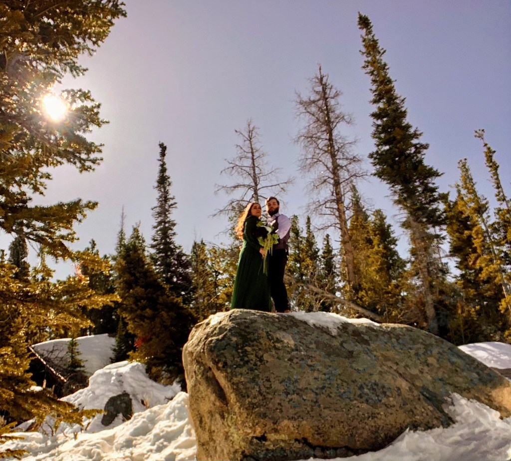 A married couple stands on a boulder overlooking a frozen lake in Rocky Mountain National Park after eloping that morning.