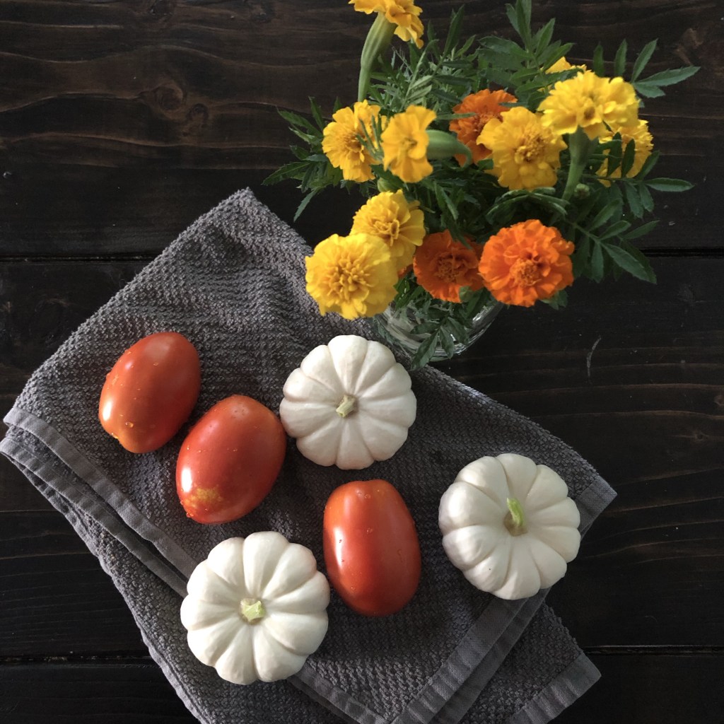 Marigolds are in a glass jar on a dining table alongside tomatoes and mini white pumpkins.