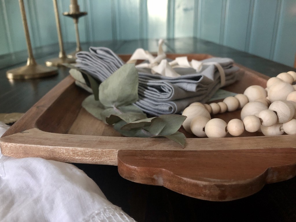 Wooden beaded garlands, eucalyptus leaves, and more sit atop a wooden serving tray on a dining table. They will be used for viewers to learn how to make a Thanksgiving tablescape.