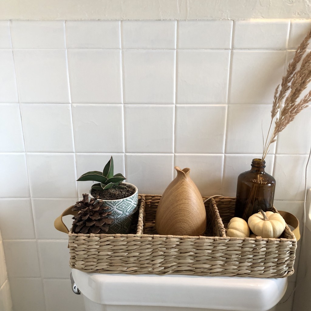 A basket near a toilet has plants, pumpkins, wheat grass, and more as added decor to a new cottage bathroom restoration project that’s just been finished. 