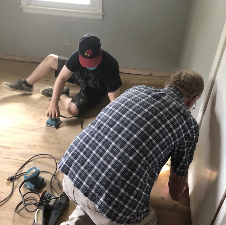 Two men work on sanding floors in a bedroom to prepare them for refinishing.