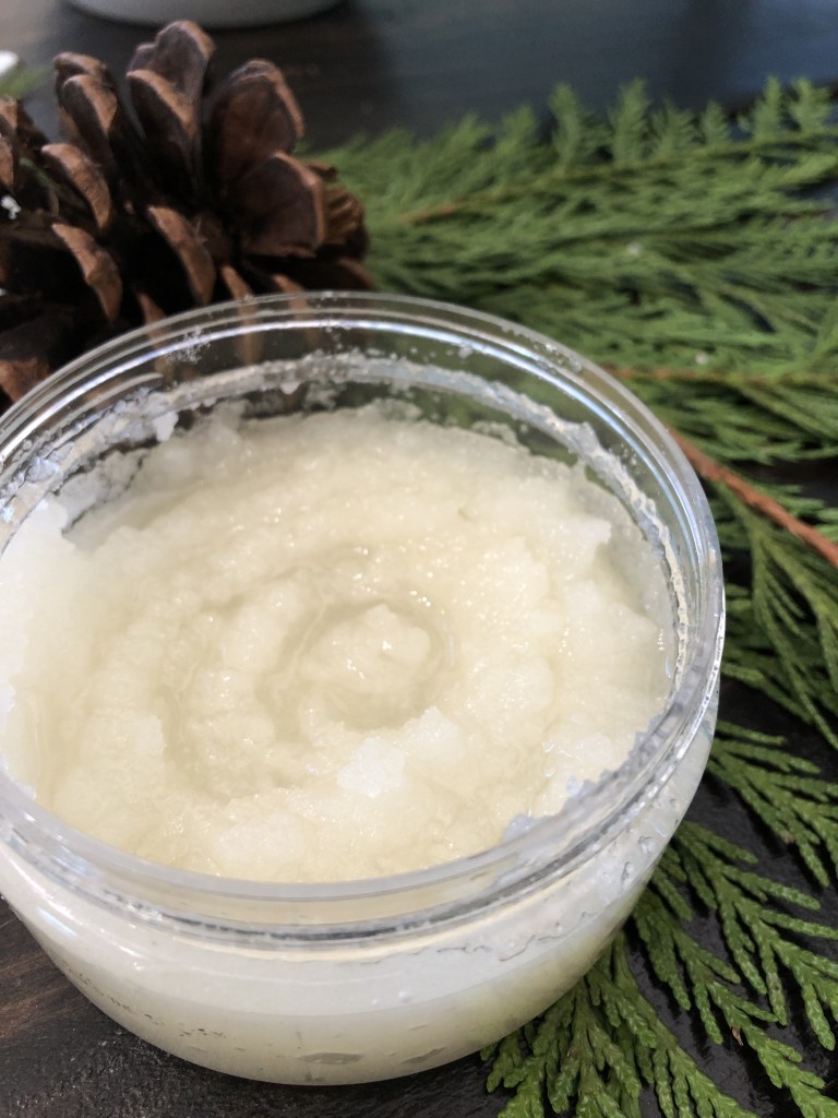 A sugar cookie body scrub smells delicious as it sots on a tabletop surrounded by fresh pine foliage.