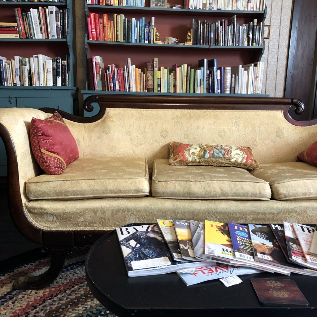 An antique couch sits in a library of a Bed and Breakfast with magazines and books scattered about.