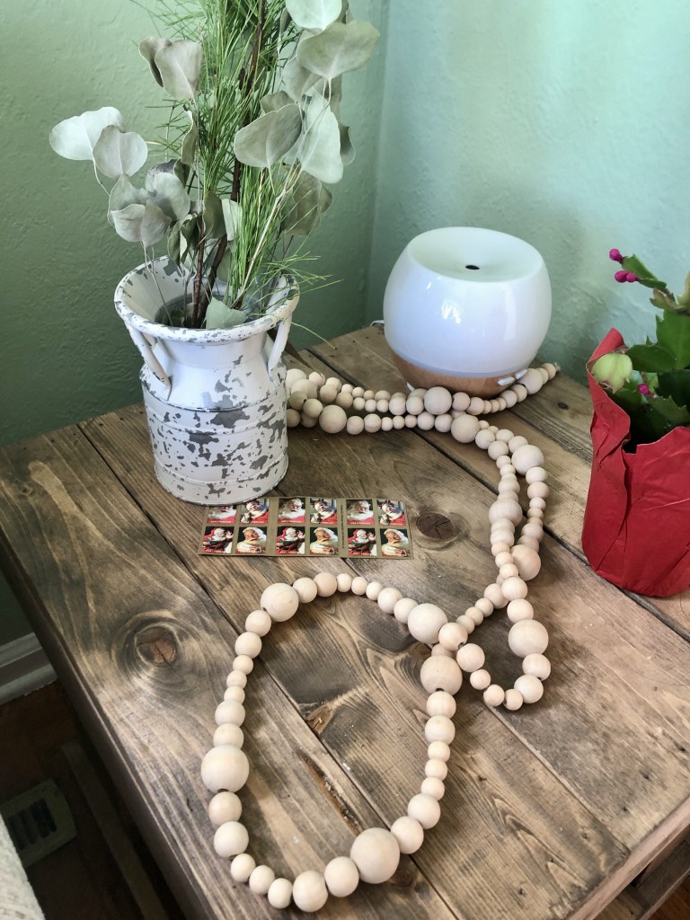 Winter cottage decor sitting on a rustic wood table. There are wooden beads, a holiday cactus, a winter green bouquet, and Santa stamps.