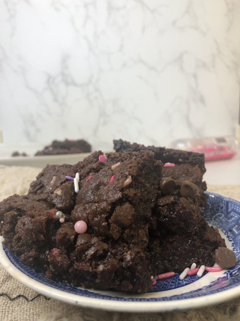 Gooey, fudge Valentine's Day Brownies have sprinkles of reds, pinks, and whites on them and are sitting on a marble table top in front of a bottle of sprinkles.
