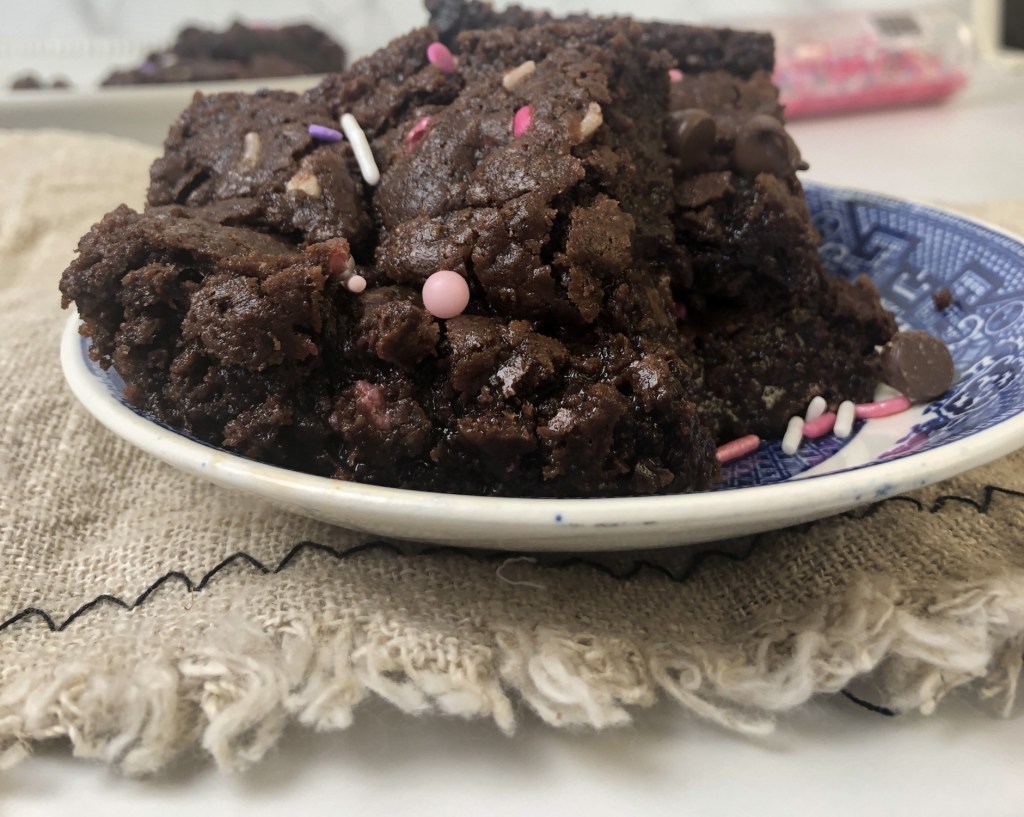 Valentine's Day Brownies sprinkled with pink and red are sitting on a marble tabletop with a linen draped under the plat it's on. Sprinkles and more brownies are in the background. 