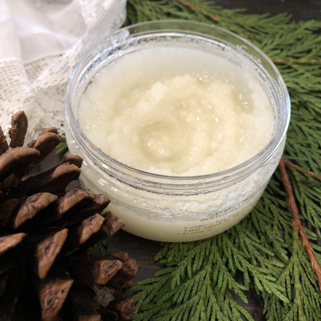 DIY sugar scrubs are great for a winter self care skin routine. A pine cone and greenery are located near a jar of sugar scrub.