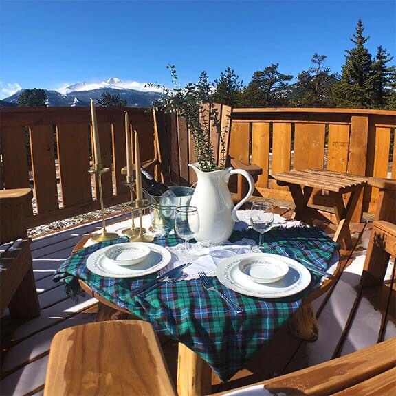 An outside deck has an array of tableware set up with snow-capped mountains showing in the background.