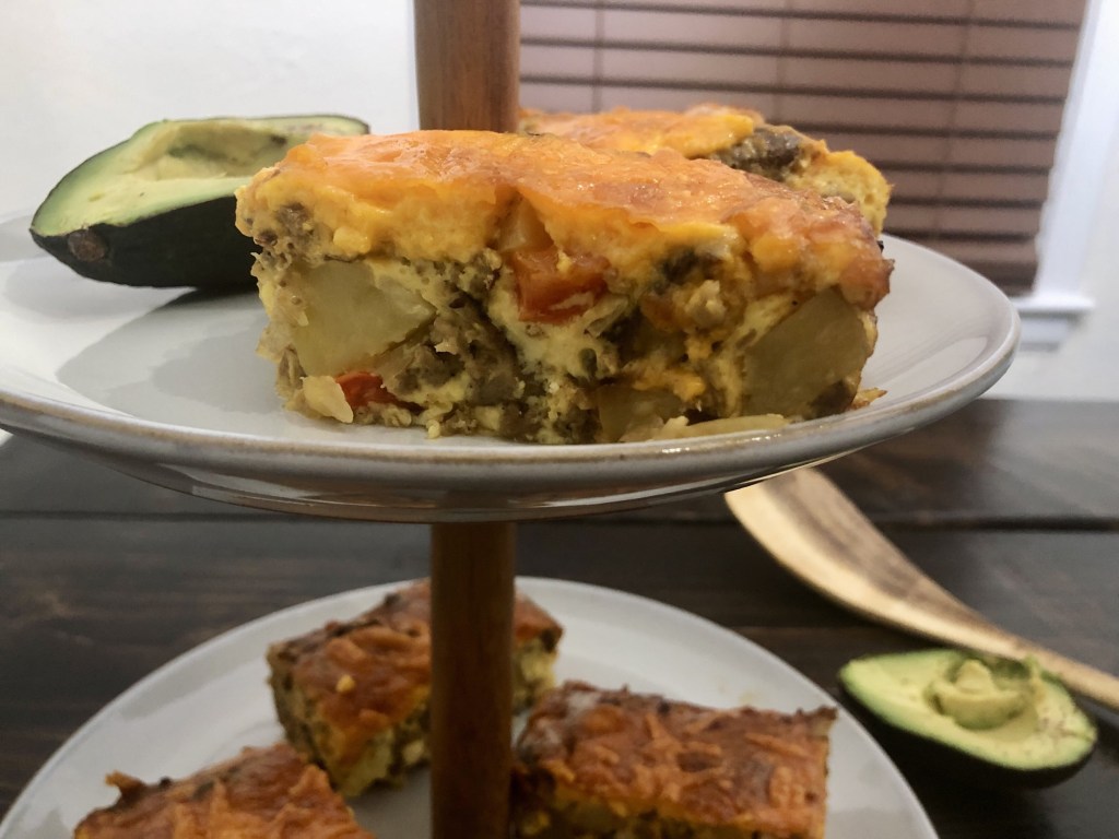 A casserole for breakfast boasts peppers, onions, vegetarian sausage, cheese and eggs as an avocado is peeped in the background.