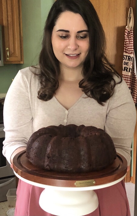 A woman brings a chocolate bundt cake into view on a cake stand.