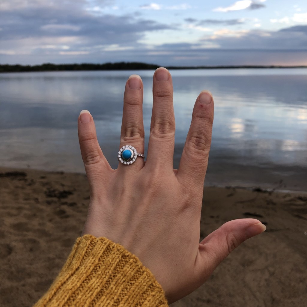 A woman shows off her turquoise engagement ring as the sun sets behind it on a lake.