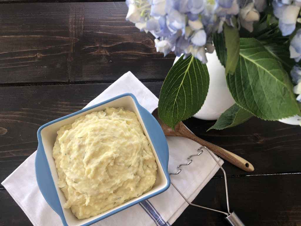 A beautiful display contains sour cream and chive mashed potatoes next to a pitcher of gorgeous blue hydrangeas.
