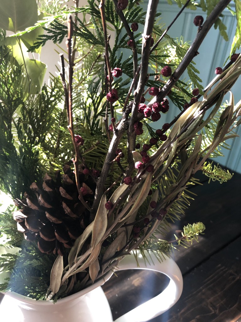 A vase full of dried branches and greenery with holly berries graces a dining table for the holiday season.