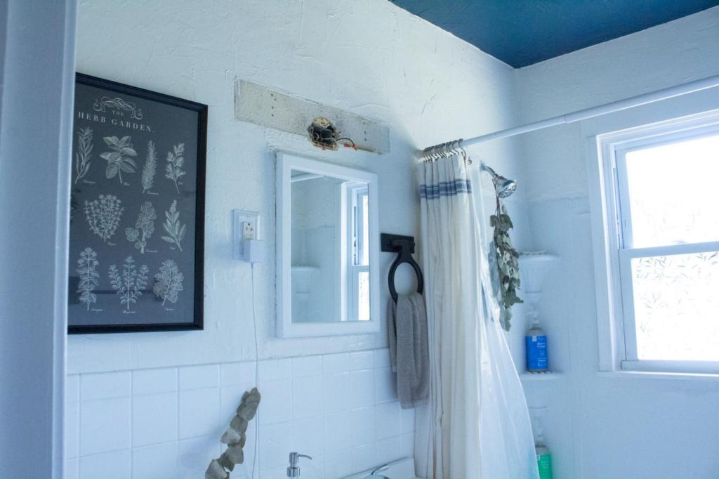 A before photo of a small cottage bathroom shows a missing light fixture, black painted wood towel racks, and cheap wall art.