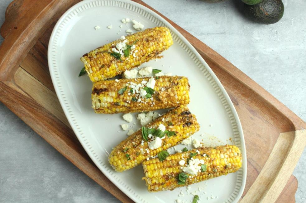 Mexican street corn is featured on a wood platter and garnished with basil and queso fresco as a vegetarian meal prep idea.