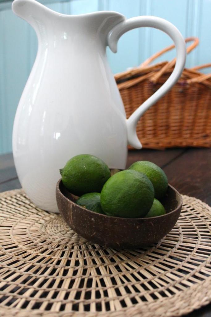 Limes in a wooden bowl are sitting in front of a pitcher and picnic basket.