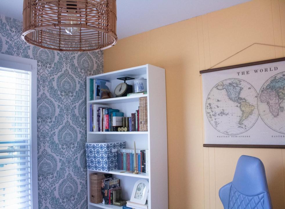 A boho style office boasts creamy orange walls, a bookcase, world map, and hanging basket light.