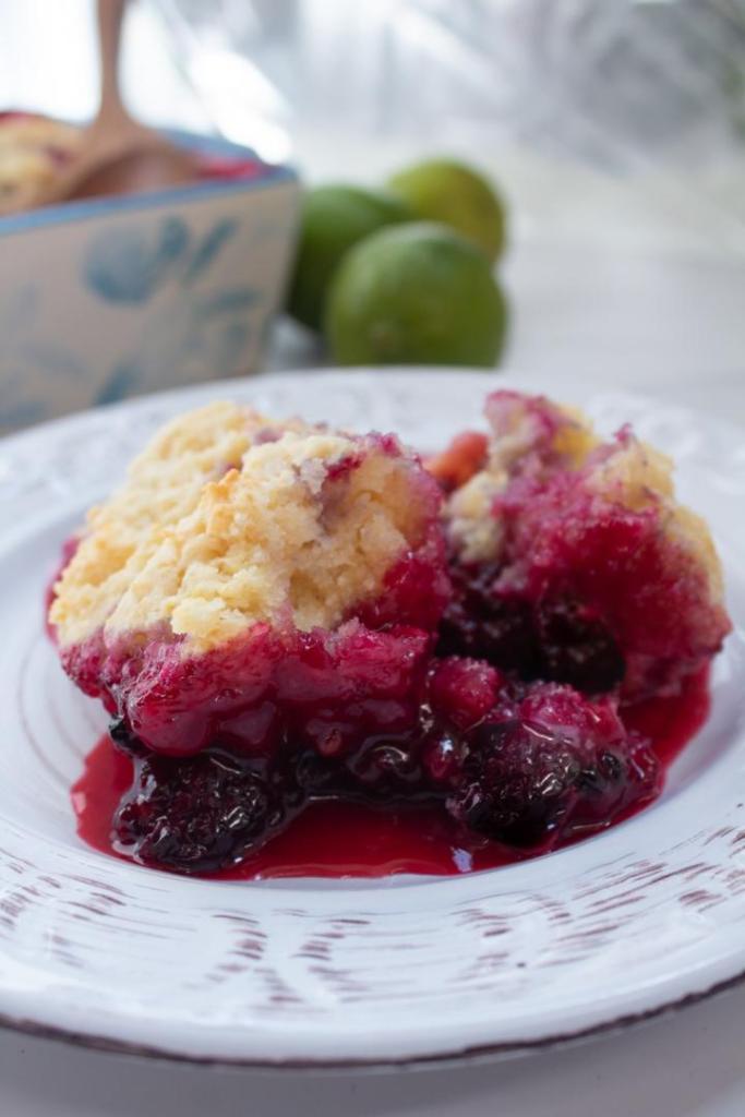 Easy Blackberry Cobbler is plated on a small dish alongside limes and a casserole dish of more cobbler behind it.