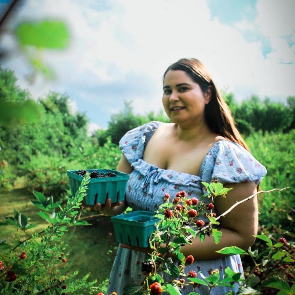 A cottagecore woman stands in an orchard holding baskets of blackberries in a flow blue dress.