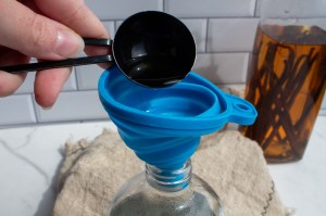A woman pours vanilla extract into a glass spray bottle over a funnel to create a DIY natural mosquito repellent.