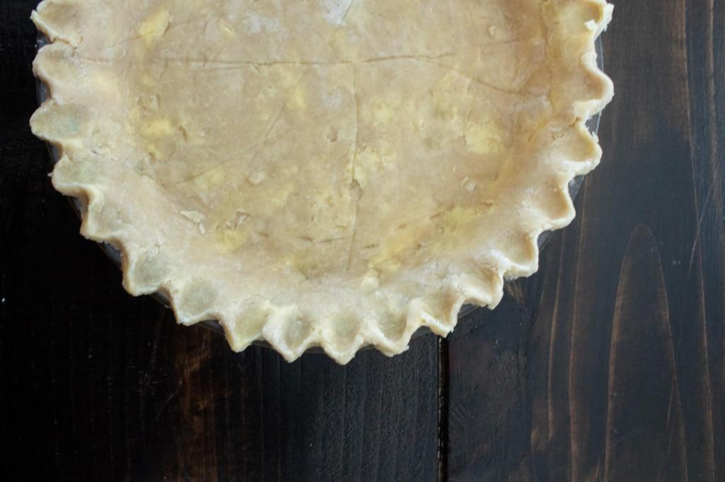 A pie crust is unbaked and sits in a pie plate on a table after the edges have been fluted.