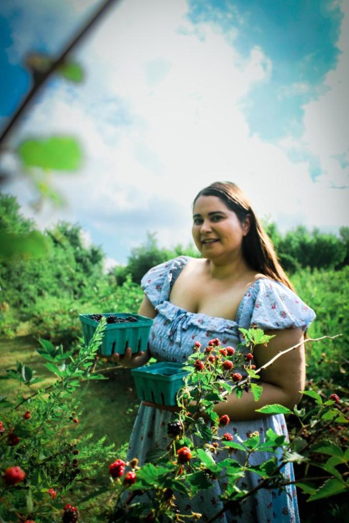 Cottagecore aesthetic is shown by a woman wearing a blue pattern floral dress in an orchard as she holds a basket of berries.