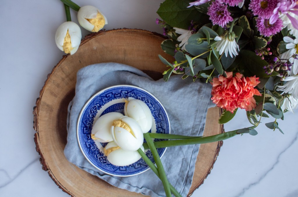 A spring tablescape is created with a hearty serving of deviled eggs and a bouquet of flowers nearby on a rustic serving board.