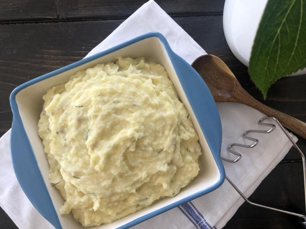 Sour cream and chive mashed potatoes are presented in a ceramic baking dish on a striped farmhouse towel.