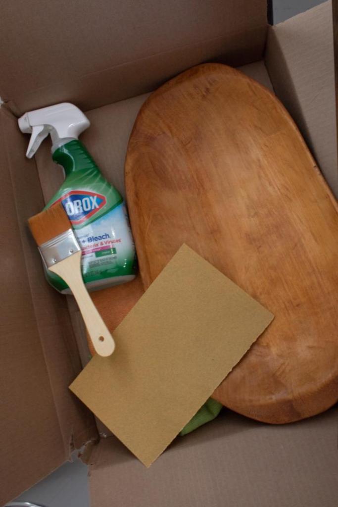 A variety of supplies are gathered into a box as a woman learns how to bleach wood decor with Clorox.