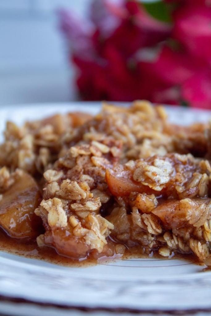 Crockpot apple crisp is the tastiest fall dessert served on a small plate with bright pink flowers next to it.